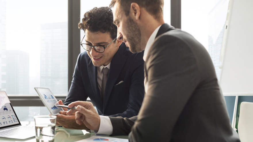 Redirection: dois homens sentados em um escritório com um computador na mesa e um tablet na mão. Eles estão conversando sobre uma aliança estratégica empresarial